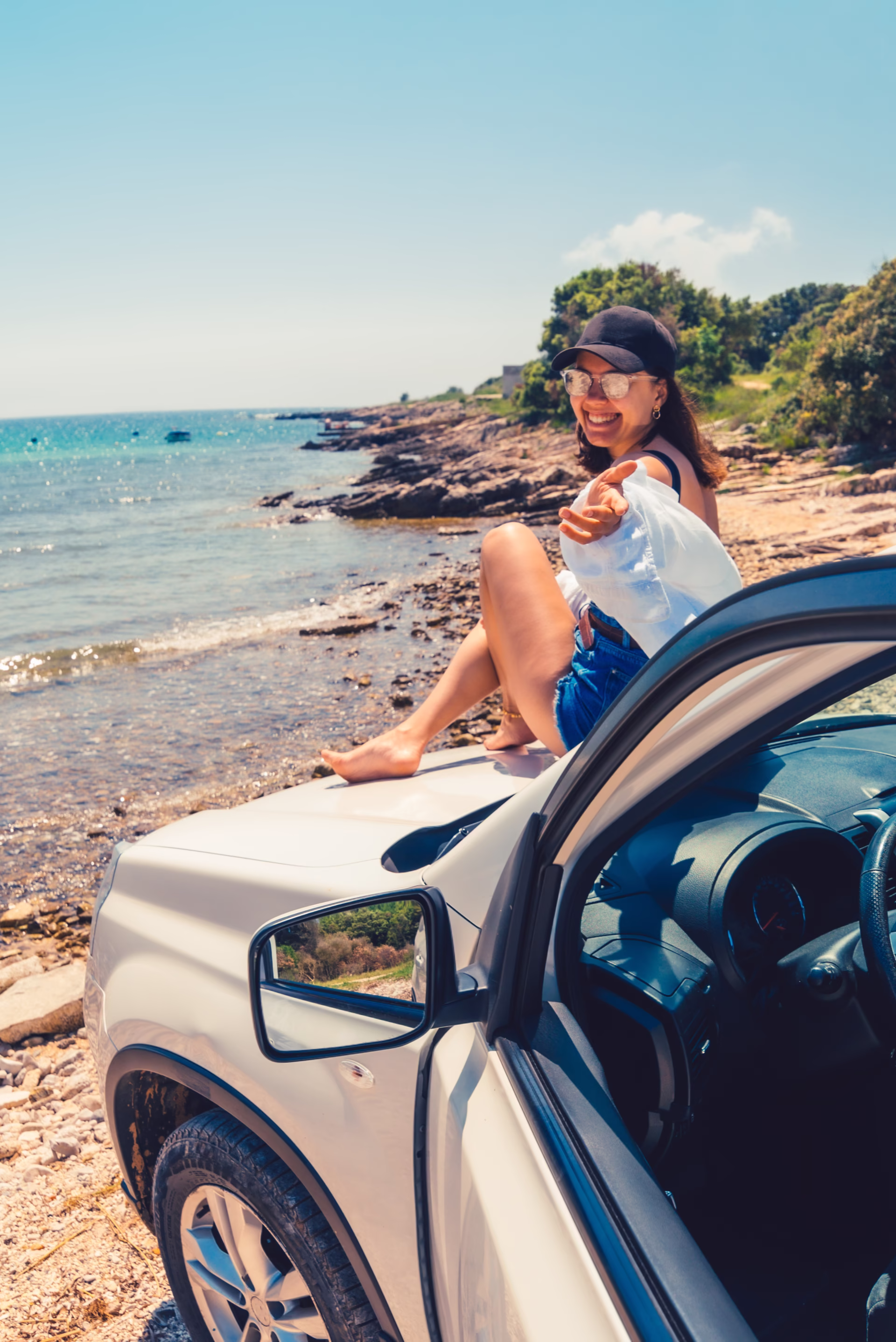 Smiling woman sitting on a car hood by the sea, representing car rental services.